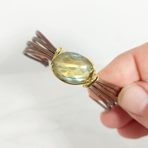 May include: A handmade bracelet featuring a faceted, oval-shaped gemstone in a gold wire setting. The gemstone has a mix of yellow, green, and blue hues. The bracelet's band is made of multiple copper-colored wires.