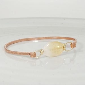 May include: A copper-colored bangle bracelet with a hammered texture. The bracelet features a light yellow, oval-shaped gemstone secured with gold-colored wire. The bracelet is displayed on a white surface.