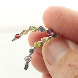 May include: A delicate, wire-wrapped ring featuring a series of colorful gemstones. The ring showcases a spectrum of colors, including red, yellow, green, blue, and purple stones. The gemstones are set in a silver-colored wire, creating a unique and artistic design.