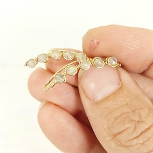 May include: A pair of gold-colored ear climbers, each featuring a series of small, round, pale gemstones. The gemstones are wire-wrapped onto the curved, ear-shaped design. The jewelry is held in a person's hand.
