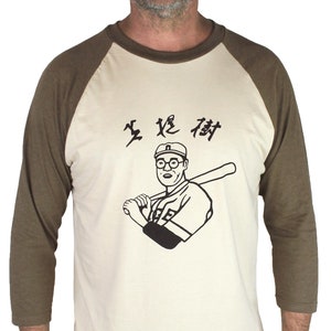 May include: A beige and brown baseball t-shirt with a black and white illustration of a man wearing glasses and a baseball cap holding a bat. The text "生 従 侍" is printed on the shirt.