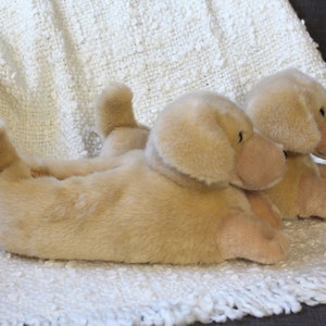 May include: Two plush golden retriever dog slippers. The slippers are lying on a white fluffy blanket.