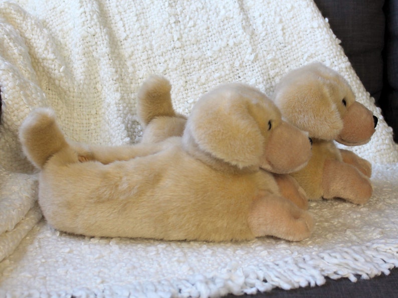 May include: Two plush golden retriever dog slippers. The slippers are lying on a white fluffy blanket.
