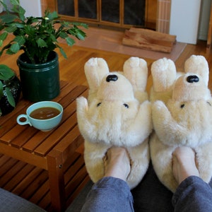 May include: A pair of beige plush dog-shaped slippers with black noses. The slippers are worn by a person sitting on a couch with a wooden coffee table in front of them.