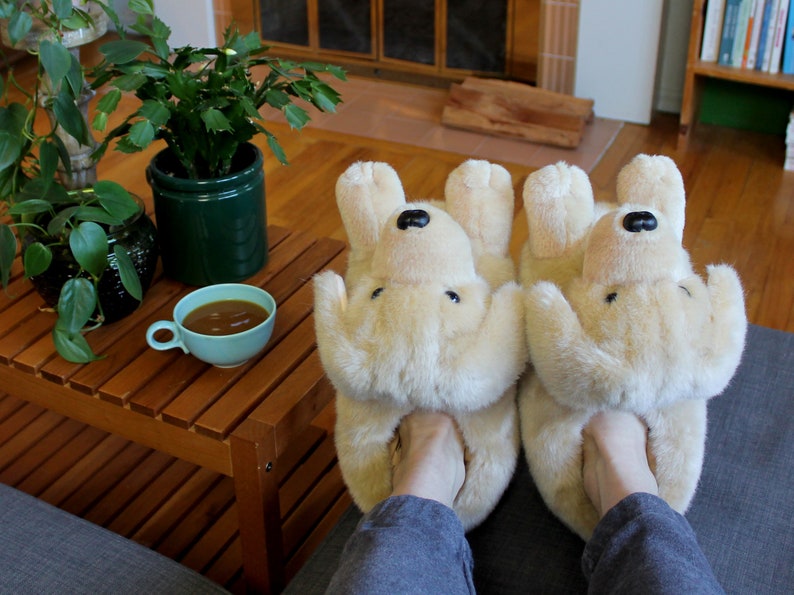 May include: A pair of beige plush dog-shaped slippers with black noses. The slippers are worn by a person sitting on a couch with a wooden coffee table in front of them.