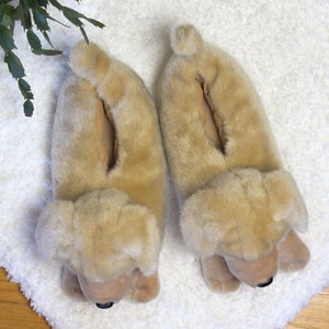 May include: A pair of beige plush dog-shaped slippers. The slippers have a furry texture and are designed to look like a dog's head and paws.