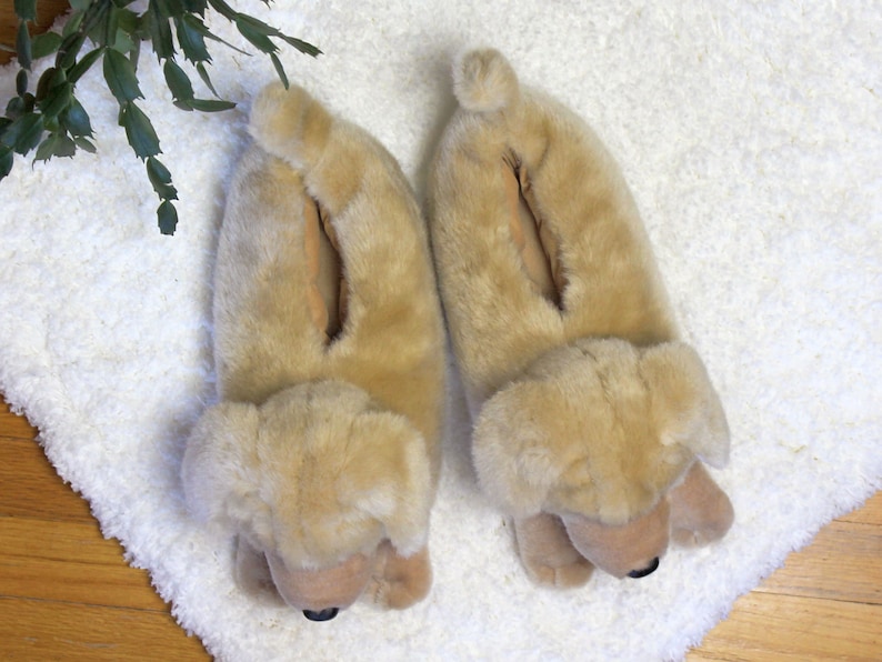 May include: A pair of beige plush dog-shaped slippers. The slippers have a furry texture and are designed to look like a dog's head and paws.