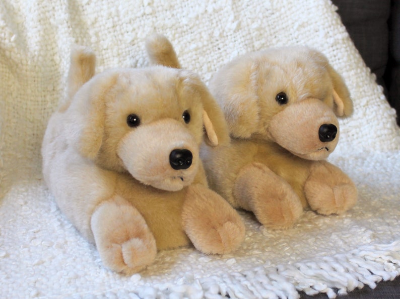 May include: A pair of beige plush dog-shaped slippers. The slippers have black eyes and noses and are sitting on a white fluffy rug.