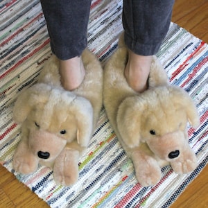 May include: A pair of tan plush slippers shaped like a dog's head. The slippers have a soft, furry texture and feature a black nose and eyes.