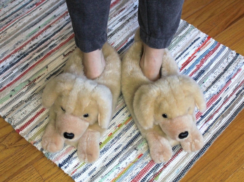 May include: A pair of tan plush slippers shaped like a dog's head. The slippers have a soft, furry texture and feature a black nose and eyes.