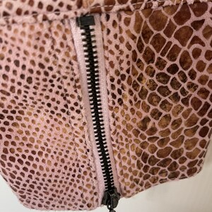 May include: Close-up of a pink and brown snakeskin-patterned fabric with a black zipper. The zipper is partially open and the zipper pull is visible.