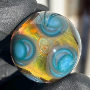 May include: A glass marble with a swirling pattern of blue, green, and yellow. The marble has a glossy finish and is spherical in shape.