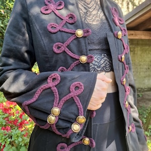 Op de afbeelding: Zwarte jas met paarse touwdetails en gouden knopen. De jas heeft een hoge kraag en decoratieve touwaccenten langs de voorkant en mouwen. Een stijlvol bovenkledingstuk.