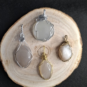 Peut inclure: Quatre pendentifs en pierres précieuses sur une surface en bois. Les pendentifs présentent diverses formes et couleurs, dont une goutte et une pierre ronde, tous enveloppés de fil d'argent ou doré. Les pierres sont gris clair et blanc cassé.