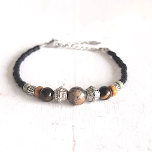 Op de afbeelding: Een kralenarmband met een zwart koord en decoratieve kralen. De armband heeft bruine, zilveren en patroon kralen, en een zilveren ketting sluiting. De armband ligt op een wit oppervlak.