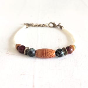 Op de afbeelding: Een armband met een centrale houten kraal met een geëtst ontwerp, geflankeerd door donkergroene en kastanjebruine kralen. De armband heeft een crèmekleurig koord en een bronzen ketting met een sluiting.