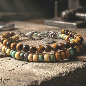 May include: A double-strand beaded bracelet featuring brown tiger eye beads and a mix of turquoise, tan, and dark wood beads. The bracelet includes a silver-toned metal lobster clasp, a short chain extender, and a small round tag engraved with Cinziadesign.