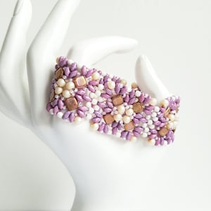 Peut inclure: Un bracelet en perles de couleur violet, blanc et or avec un motif géométrique. Le bracelet est fait de petites perles de rocaille et de perles carrées plus grandes.