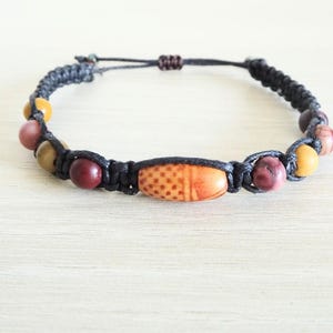 Op de afbeelding: Een macramé armband met een centrale cilindrische kraal met stippenpatroon, geflankeerd door ronde kralen in tinten bordeauxrood, mosterdgeel en bruin. De armband is gemaakt van zwart koord en heeft een verstelbare schuifknoopsluiting.