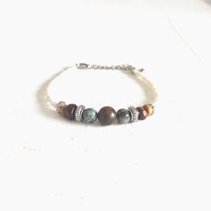 Op de afbeelding: Een armband met een crèmekleurig koord en een zilverkleurige ketting sluiting. Het heeft natuurstenen kralen in bruin, groen en grijs, met zilverkleurige afstandhouders. De armband is een delicaat sieraad, ongeveer 18 cm lang, voor dagelijks gebruik.