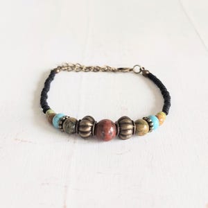 Op de afbeelding: Een kralenarmband met een zwart koord en kleurrijke kralen. De armband bevat turquoise, bruine en goudkleurige kralen, een centrale roodbruine steen en antiek messingkleurige metalen kralen. De armband is ongeveer 17,78 cm lang.