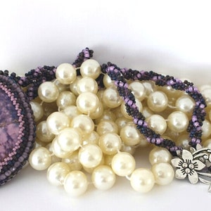 May include: A purple and brown beaded necklace with a large purple stone pendant. The necklace is layered on top of a strand of white pearls.