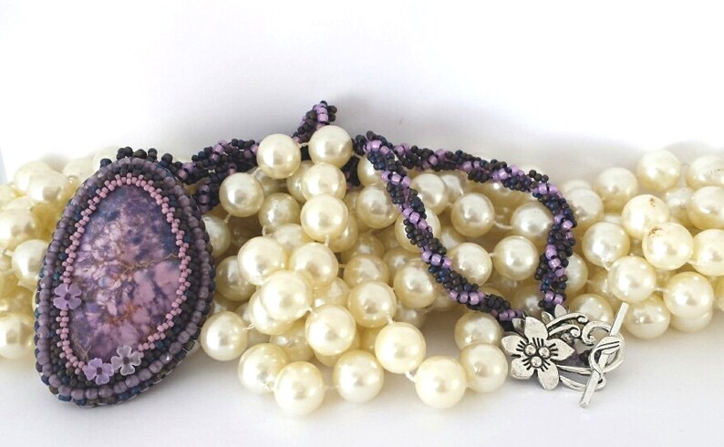 May include: A purple and brown beaded necklace with a large purple stone pendant. The necklace is layered on top of a strand of white pearls.