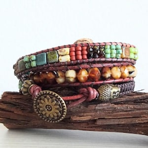 May include: A multi-strand wrap bracelet featuring brown leather cords, turquoise, red, and green square beads, and a row of faceted amber-colored beads. The bracelet is secured with a round, antique-style brass button closure with a floral design.