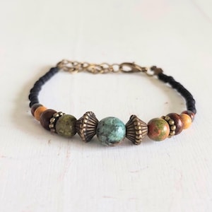 Op de afbeelding: Een kralenarmband met een zwart koord, houten kralen in bruin- en oranjetinten en decoratieve metalen accenten. De armband heeft een centrale turquoise steen en een goudkleurige ketting voor verstelbare maatvoering.