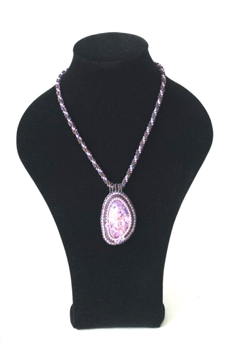 May include: A purple beaded necklace with a large, oval-shaped pendant. The pendant is made of purple beads and has a textured, iridescent finish.