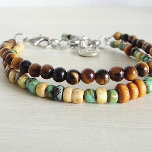 Op de afbeelding: Een dubbelstrengs armband met één rij bruine tijgeroogkralen en een tweede rij van afwisselend turquoise, crème en lichtbruine houten kralen, afgewerkt met een zilverkleurige metalen karabijnsluiting en een kleine ronde bedel.