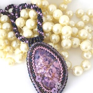 May include: A purple and pink stone pendant with a beaded border and small pink flower accents. The pendant is hanging from a strand of white pearls.