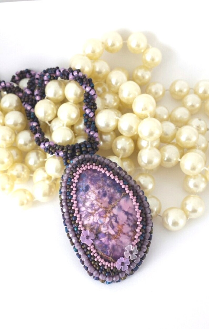 May include: A purple and pink stone pendant with a beaded border and small pink flower accents. The pendant is hanging from a strand of white pearls.