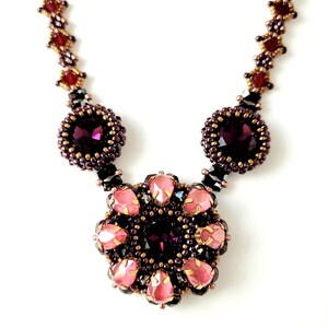 Op de afbeelding: Een kralenketting met een roze en paarse bloempendant. De ketting is gemaakt met kleine, gefacetteerde kralen in tinten bruin, rood en zwart. De hanger heeft een centrale paarse kristal omgeven door roze en paarse kralen.