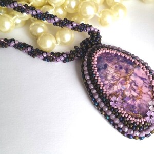 May include: A purple and brown beaded necklace with a large purple and gold stone pendant. The pendant is surrounded by smaller beads and has three small pink flowers.