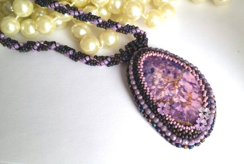 May include: A purple and brown beaded necklace with a large purple and gold stone pendant. The pendant is surrounded by smaller beads and has three small pink flowers.