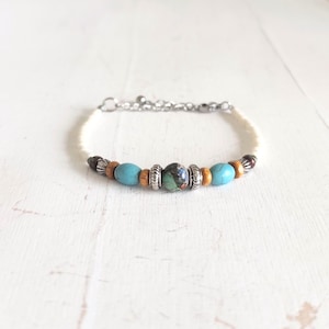 Op de afbeelding: Een armband met turquoise, houten en patroon kralen. De armband heeft een zilveren ketting en sluiting. De kralen zijn geregen op een crèmekleurig koord. De armband wordt gepresenteerd op een wit oppervlak.