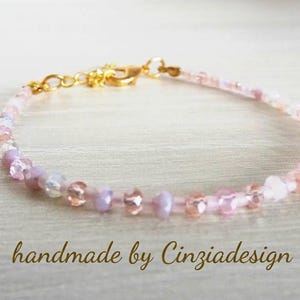 Op de afbeelding: Een delicate armband met kleine, gefacetteerde kralen in tinten lichtroze, lavendel en helder kristal. De armband is voorzien van een goudkleurige metalen karabijnsluiting en een korte verlengketting. De tekst handmade by Cinziadesign is zichtbaar.