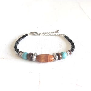 Op de afbeelding: Een kralenarmband met zwarte, bruine en turquoise kralen, en een houten tonvormige kraal. De armband heeft een zilverkleurige ketting en sluiting. Het ontwerp is casual en bohemien.