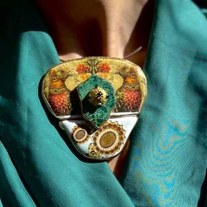 Op de afbeelding: Een grote, driehoekige, keramische hangerketting met een groen, goud en oranje ontwerp. De hanger wordt gedragen door een persoon met een groene top.