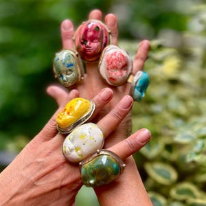 May include: A collection of six rings featuring sculpted doll face motifs in various colors including pink, white, yellow, blue, and green. Each face is set in a metallic band, showcasing distinct painted details and glossy finishes on each piece.