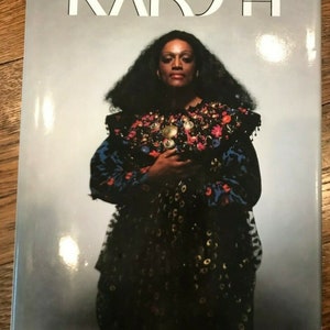 May include: A book titled "Karsh" featuring a black and white photograph of a woman with dark hair wearing a black and blue patterned dress with gold accents.