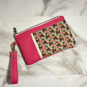 May include: A hot pink clutch with a wrist strap. The clutch features a zipper closure and a patterned design of pineapples and green leaves on a pink background. The clutch has a gold-colored metal clasp and zipper pull.