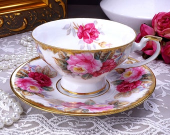 May include: A white teacup and saucer set with a gold rim and floral pattern. The floral pattern features pink roses and green leaves. The set is on a white lace tablecloth.