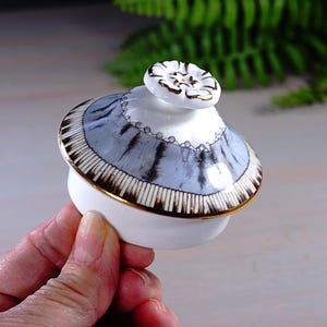May include: A white ceramic lid with a blue and black striped pattern and gold trim. The lid has a small, white floral knob with gold accents. The lid is likely for a decorative jar or container.