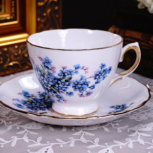 May include: A white teacup and saucer with a floral pattern of blue forget-me-nots. The cup and saucer have a gold trim.