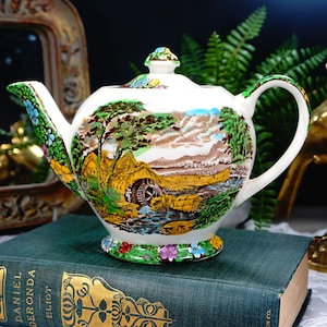 May include: A white teapot with a green, brown, and yellow landscape scene. The teapot has a floral design around the base and a spout and handle.