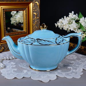 May include: A light blue ceramic teapot with gold trim and a matching lid. The teapot has a fluted design and is decorated with gold leaf patterns. The teapot is sitting on a white lace doily. A gold framed picture and white flowers are in the background.