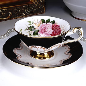 May include: A black teacup and saucer set with a floral rose design. The teacup features a black exterior with gold trim and a white interior with pink roses. The saucer has a black and white scalloped edge with gold accents.