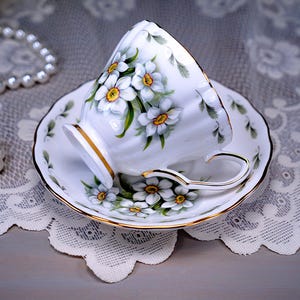 May include: White teacup and saucer set with a floral design featuring white flowers with yellow centers and green leaves. The teacup and saucer have gold trim. A pearl necklace is in the background.